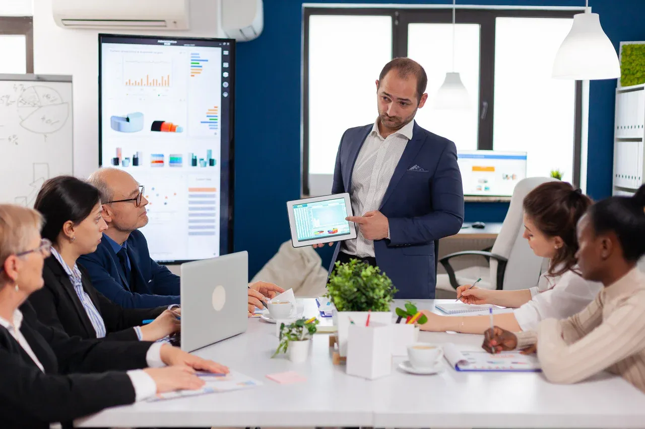 Presenter sharing charts on a tablet with colleagues around a conference table